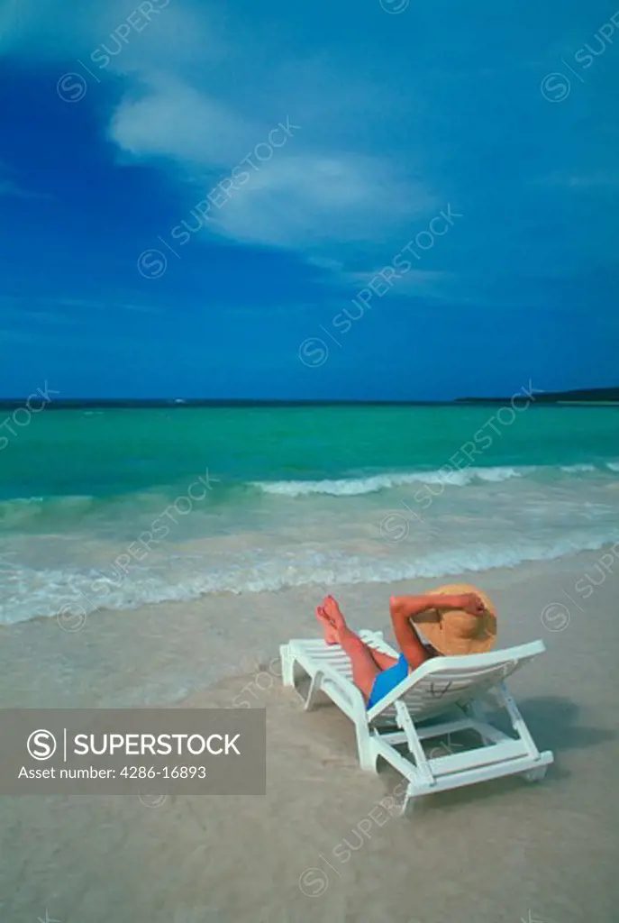 A woman wearing a wide brimmed hat looks out over the ocean while laying on a white lounge chair at the edge of the water.