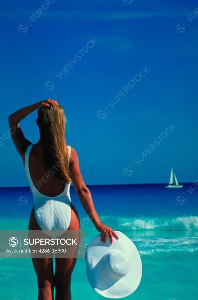 A woman wearing a white bathing suit and holding a white wide brimmed hat looking out across crystal blue water at a sail boat in the distance.