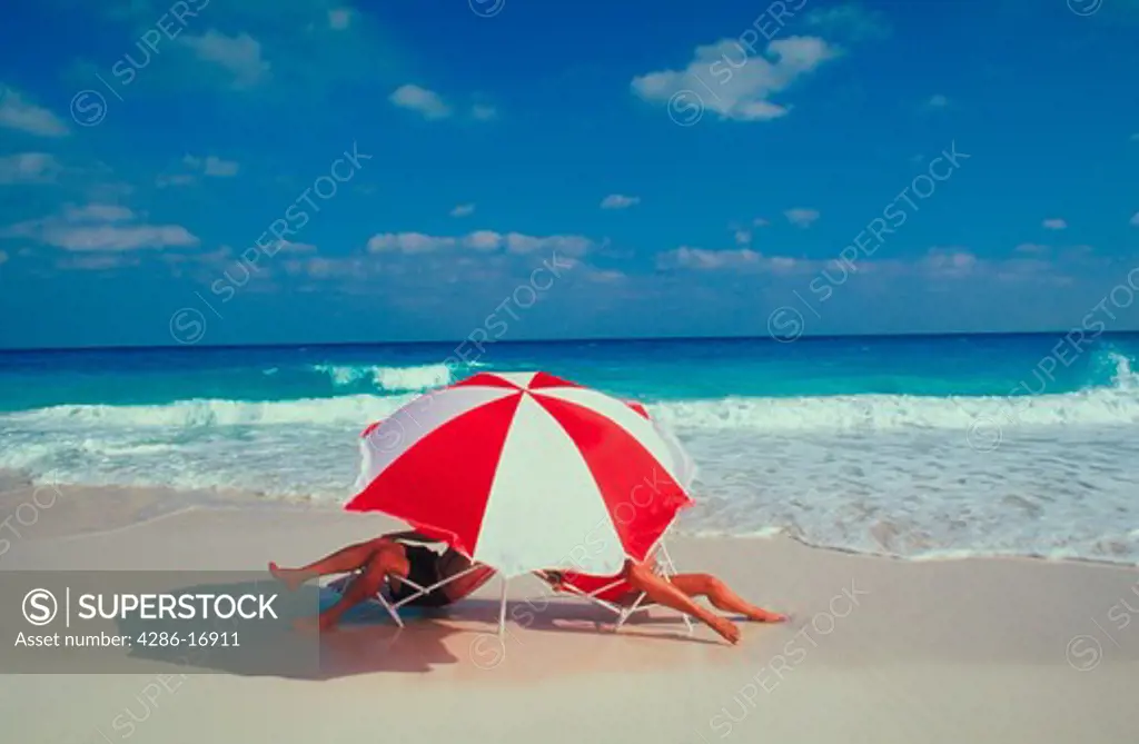 A man and a woman relaxing in beach chairs at the waters edge beneath a red and white umbrella.