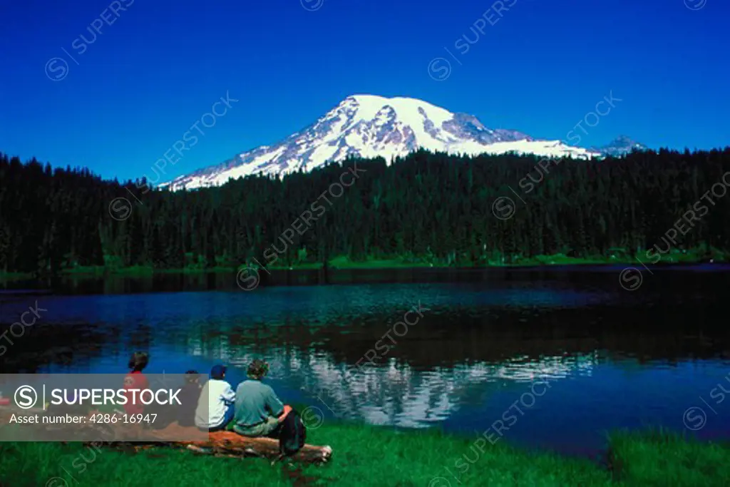 A family resting on a fallen log at Reflection Lake with a snow capped Mount Rainier, Washington in the distance.