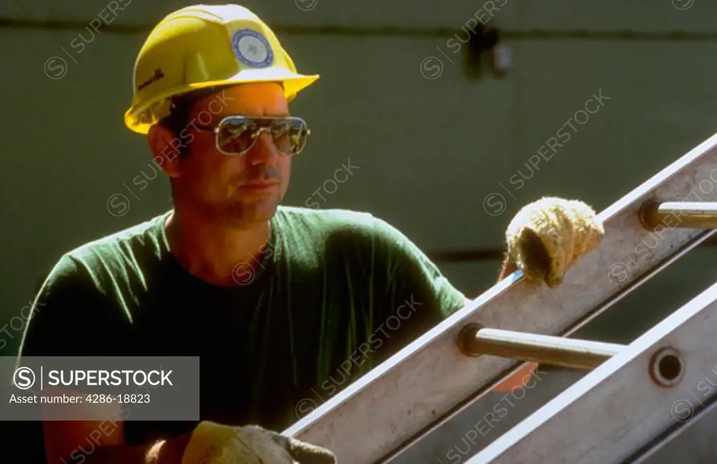 Portrait of a construction worker weaing gloves and a hardhat standing next to a ladder.