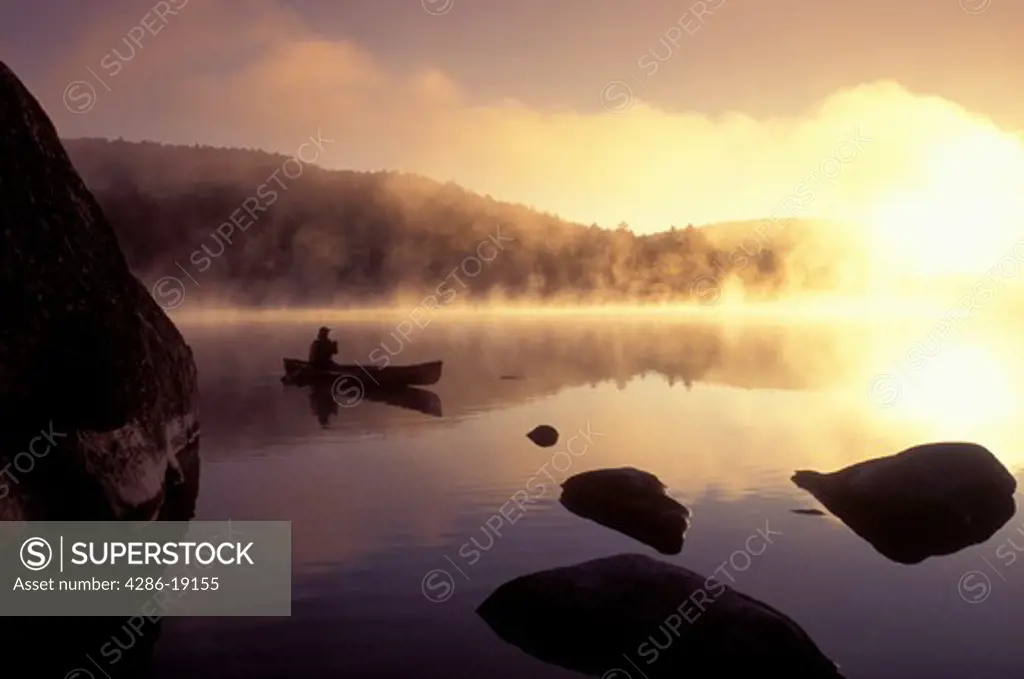 Vermont, canoe, canoeing, Woman paddles canoe in the early morning fog, mist on Kettle Pond atsunrise, sunset in Groton State Forest. Mad River Canoe Products.