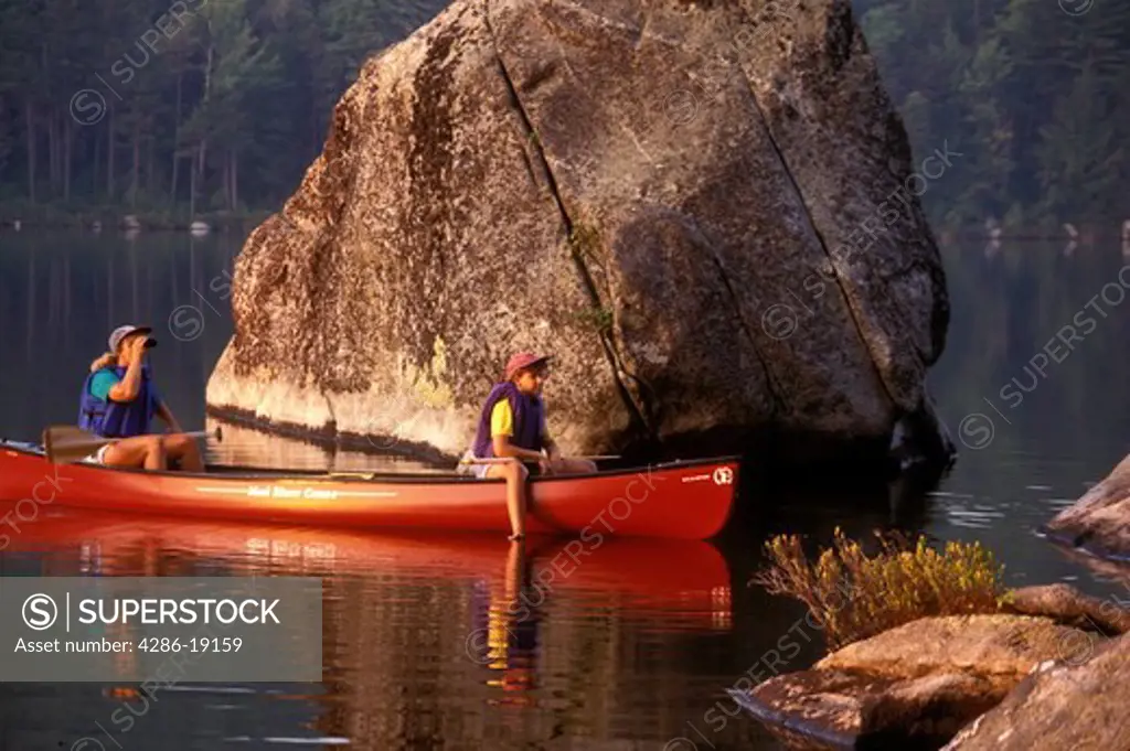 Vermont, canoe, canoeing, Mother and daughter sitting together in canoe on Kettle Pond next to a huge boulder in Groton State Forest. Daughter relaxing with feet in water. Mother looking through binoculars. Mad River Canoe Products.