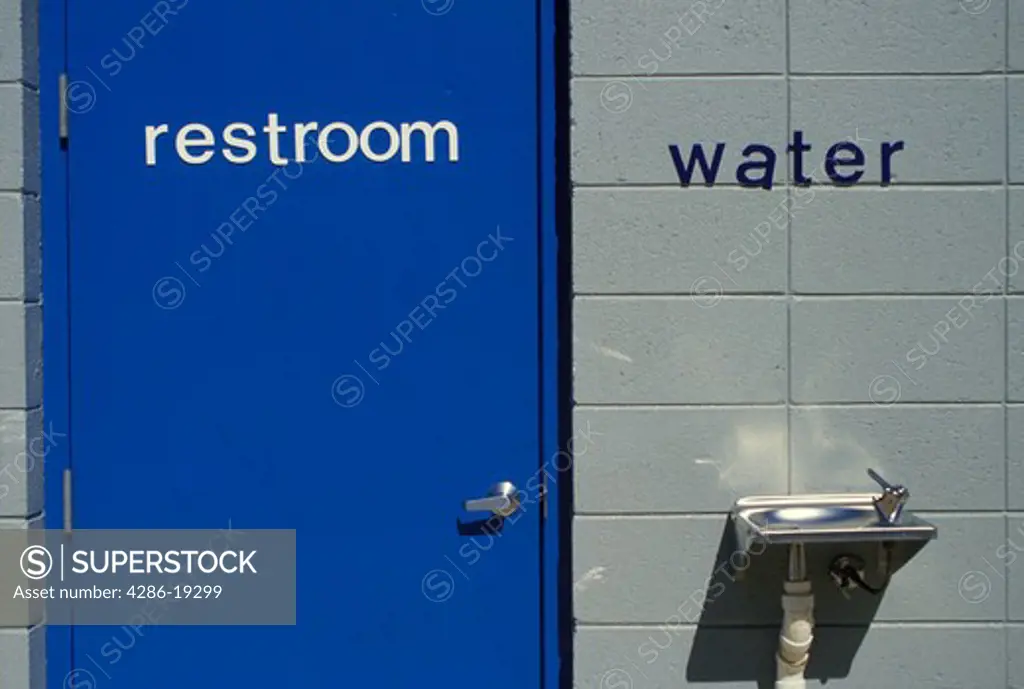 restroom, water fountain, Blue door of a restroom next to a water fountain hanging on a gray wall. The words (restroom, water) are on the (door, wall) .