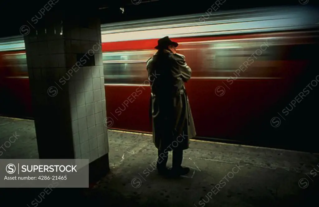 Businessman wearing trenchcoat waiting for subway train on platform, while another train speeds by.
