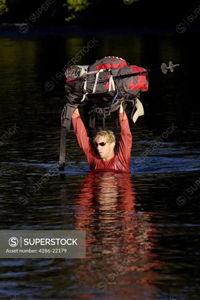 A man carrying a backpack over his head while crossing a river.
