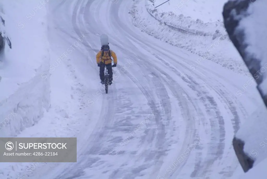 Man riding a mountain bike with a snowboard on his back in a snowstorm.