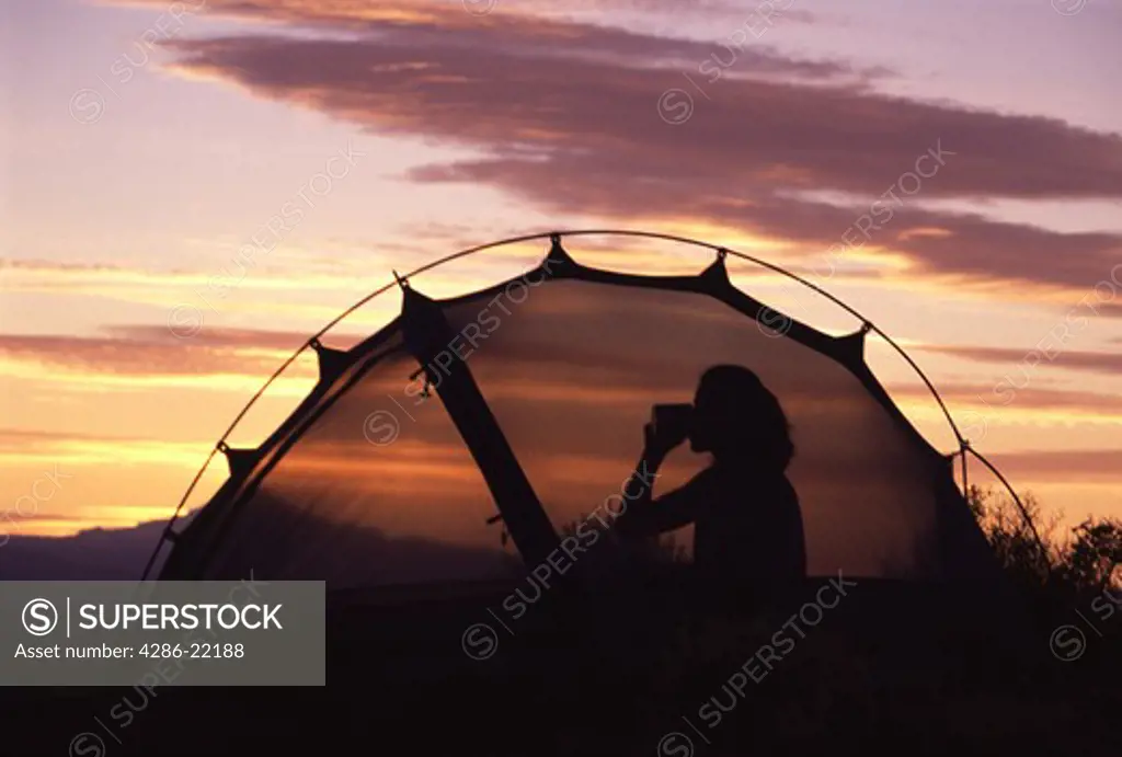 A silhouette of a woman in her tent drinking during a sunset at Smith Rock, OR.