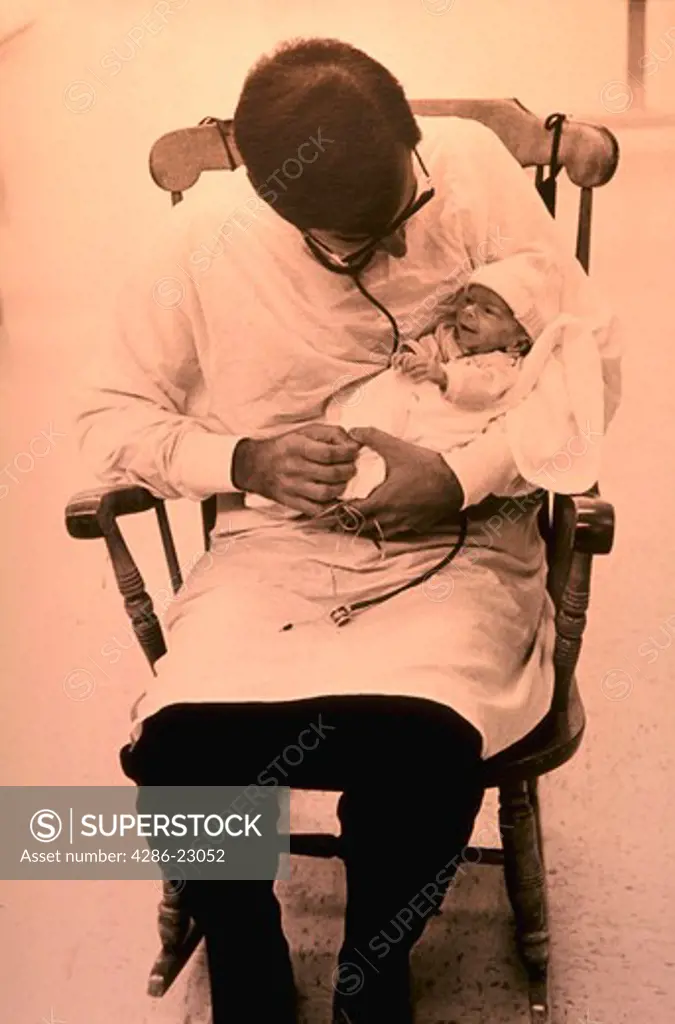 Doctor wearing white lab coat and sitting in rocking chair while holding premature baby in his lap.  The baby appears to be having a conversation with doctor.