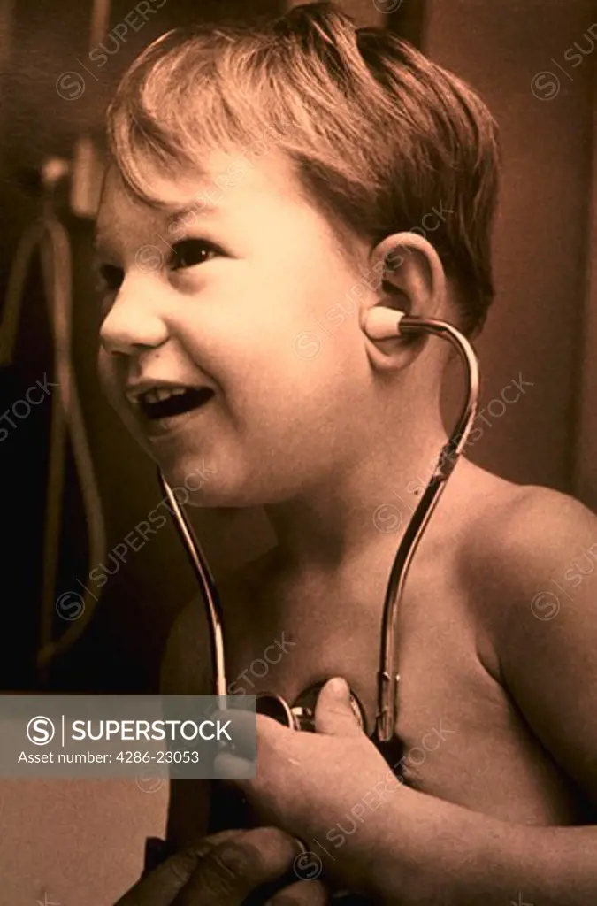 B/W  5-6yo white boy in hospital listens to his heartbeat while holding stethoscope.