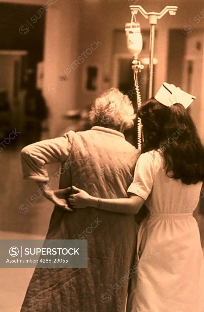 B/W Elderly female patient walks down the hall with an IV pole as nurse supports her. Nice hands backs to camera.