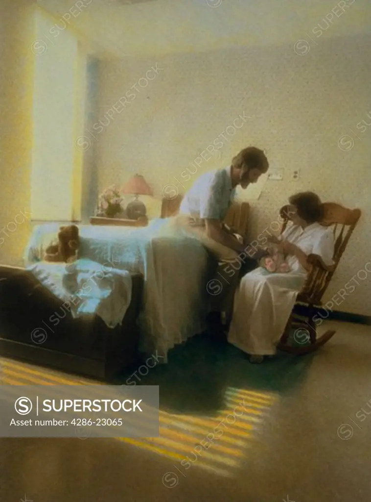 Hand colored image of man wife and newborn in birthing room with sunlight on floor.