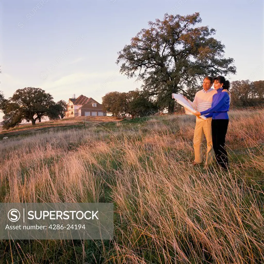 Middle-age couple stand in a field gazing into the sunset as they hold a set of blueprints. Nearby a large house is being built.