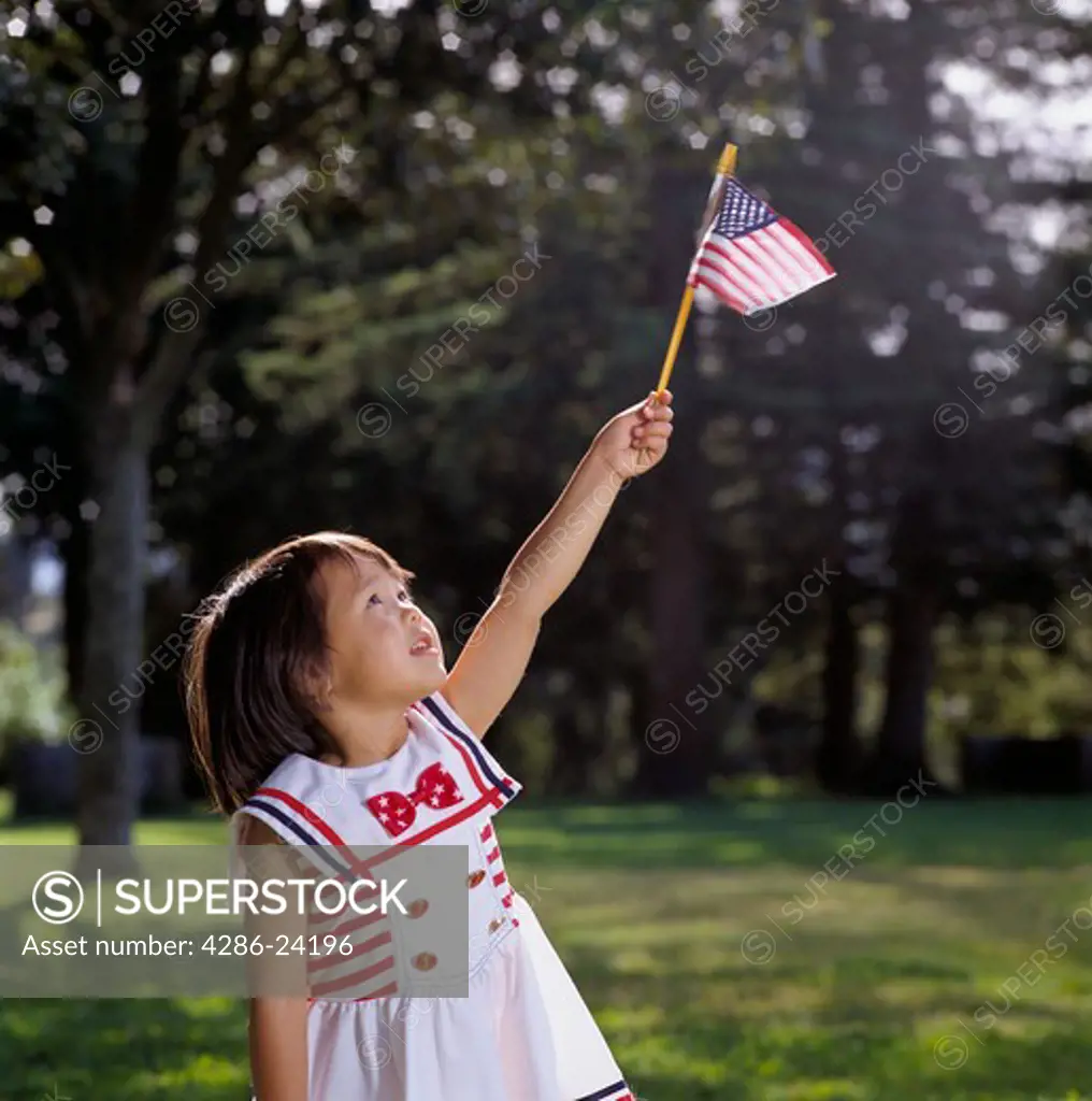 Young girl outdoors wearing a white dress with red bow holds a small U.S. flag aloft in her hand and looks skyward.