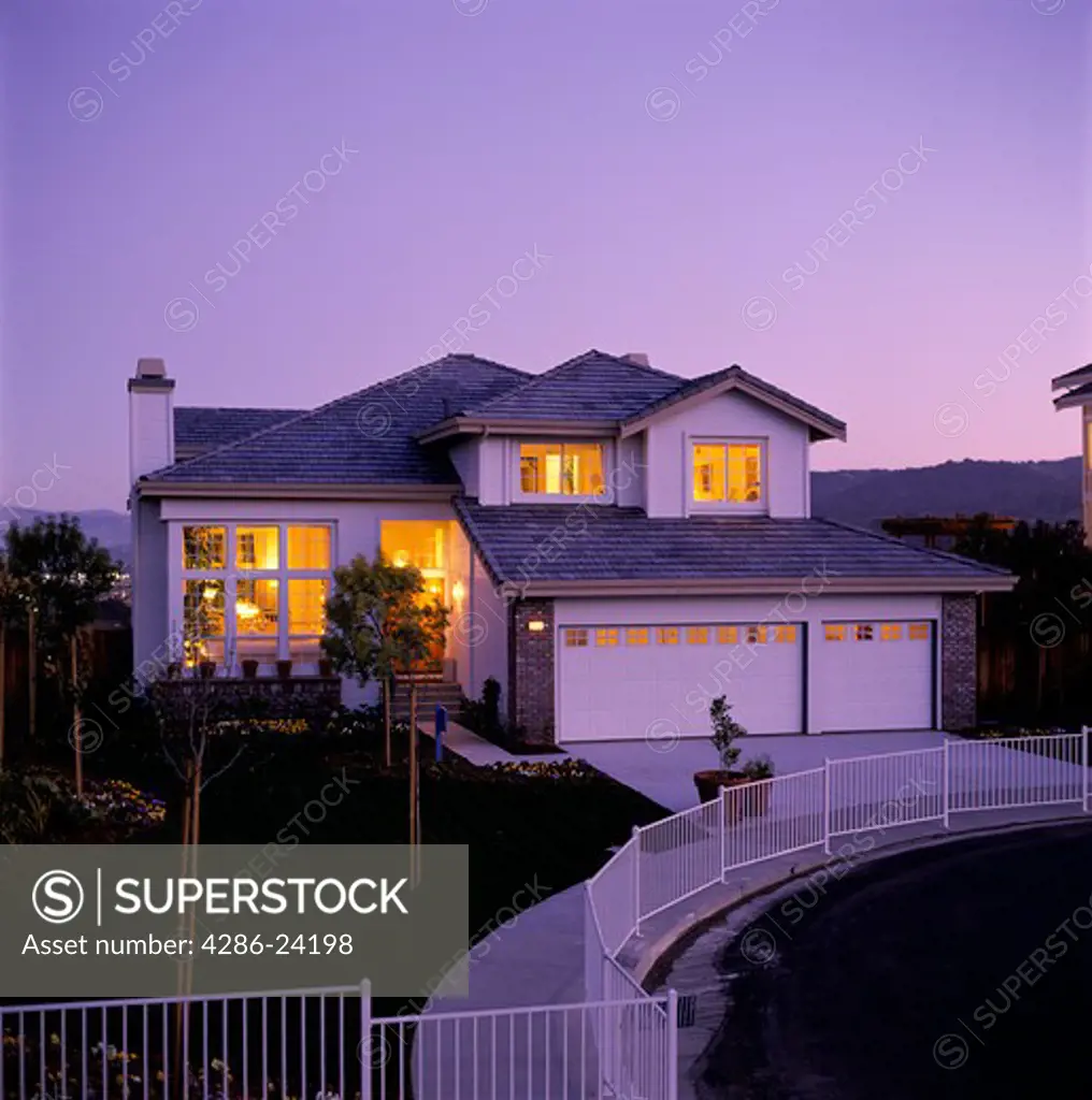 Warm yellow light shines out the windows of a suburban house in pink evening twilight. A white iron fence encloses a sidewalk. 