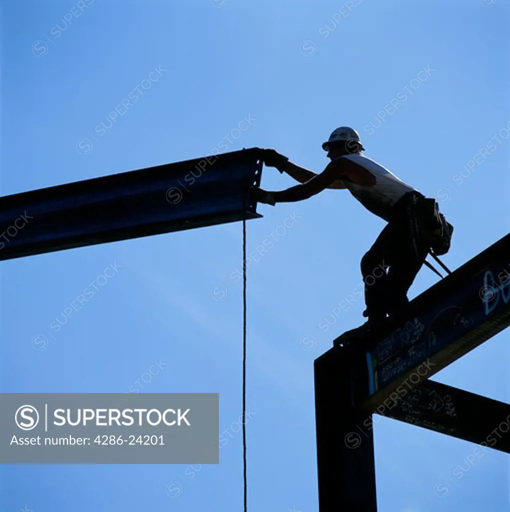 Silhoutted ironworker balancing on steel framework reaches for a girder to manuver it into place on buliding construction site.