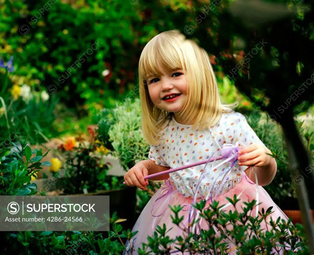 A blonde three year old girl wearing a fairy costume and holding a magic wand stands in a garden.