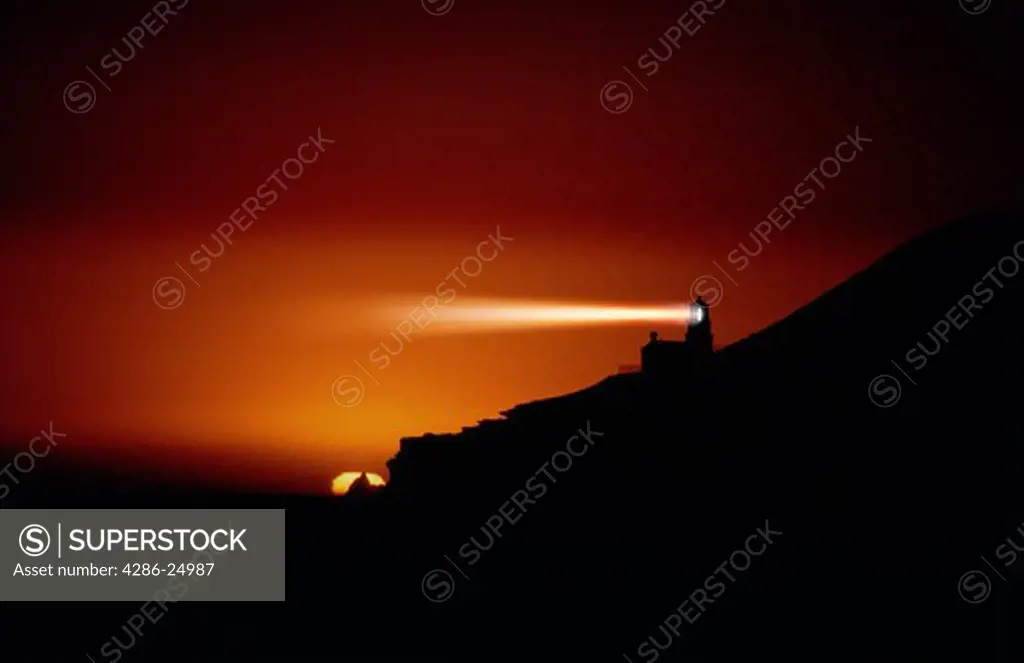 Light beams radiate from Point Conception lighthouse against orange sky just after sunset, California.