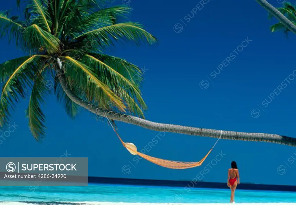 Woman wearing a red swimsuit standing next to a hammock hanging from a palm tree on a white sandy beach, Maldives Islands.