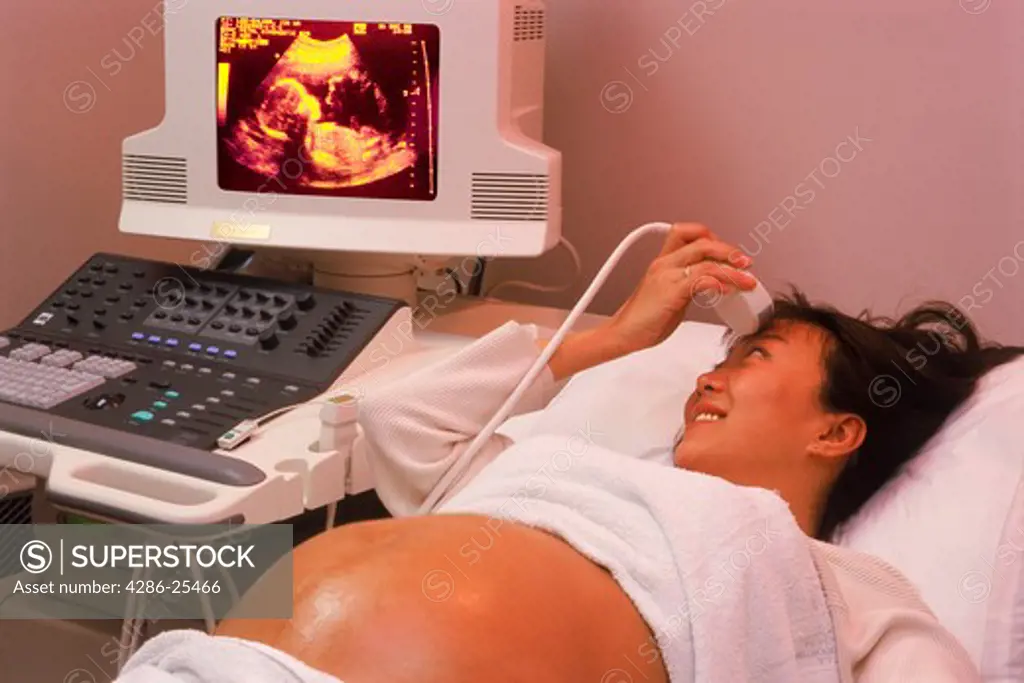 Pregnant woman holding ultrasound probe to forehead while looking at her fetus on screen