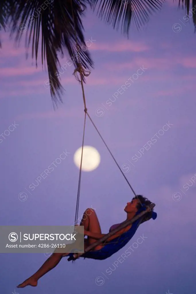 Woman in swing hanging from palm tree with full moon