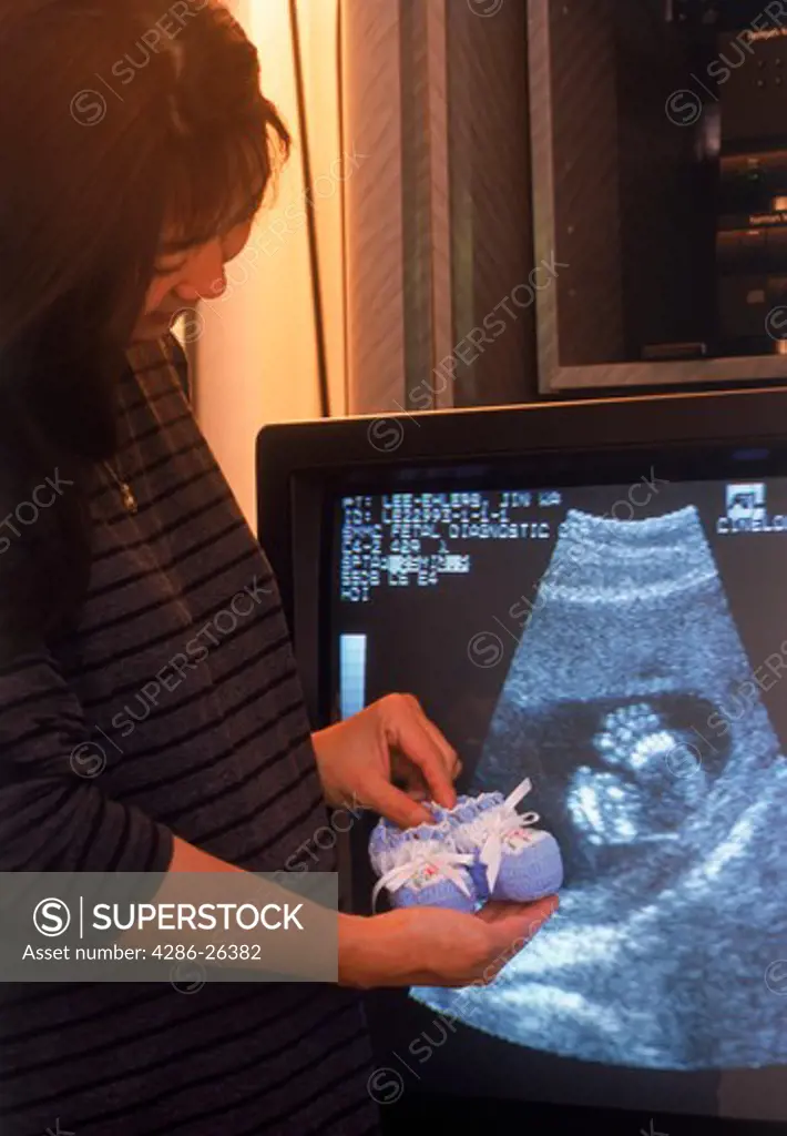 Pregnant mother with baby booties next to feet of her fetus shown on ultrasound video 