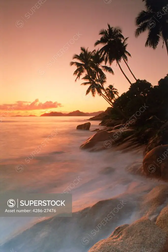 Palm trees and waves over granite rock formations on La Digue Island at sunset