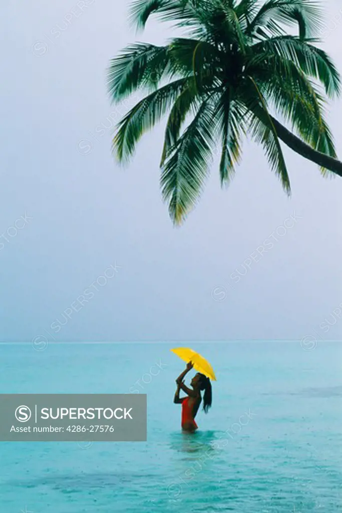 Woman standing under palm tree and yellow umbrella in warm tropical rain