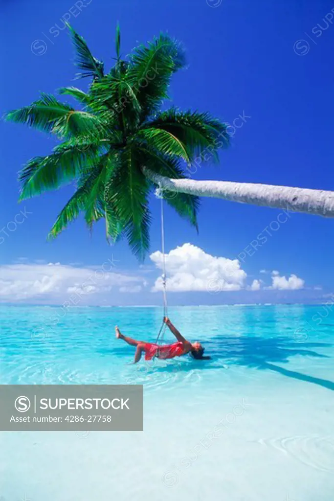 Woman laying in swing over aqua waters under palm tree and cloud on Meeru Island in Maldives