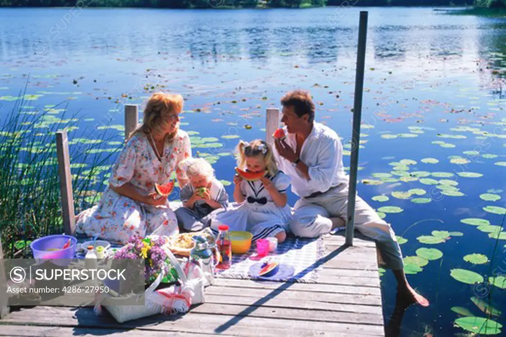 Family of four with picnic basket on lakeside pier during Swedish summer