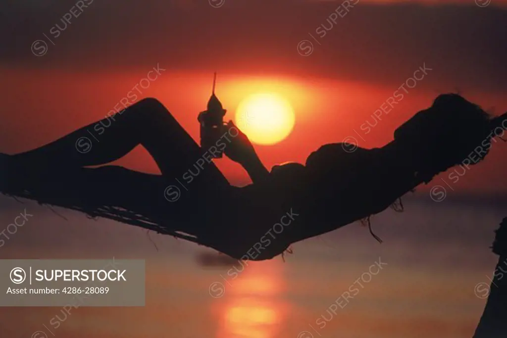 Woman in hammock with drink in hand silhouetted at sunset