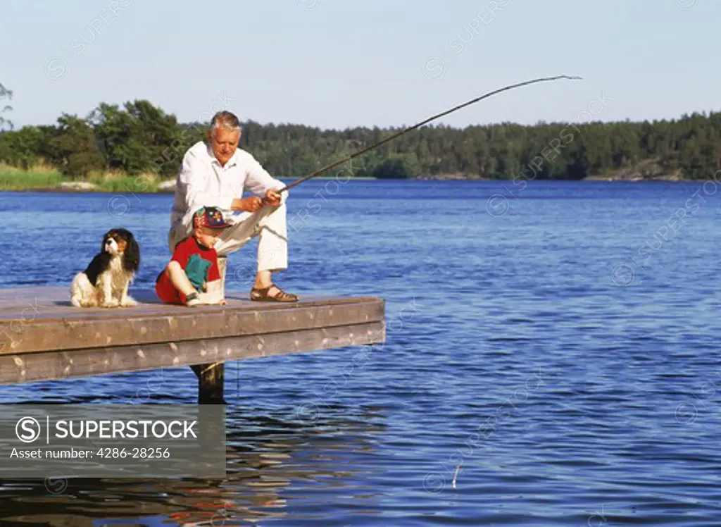 Grandad fishing with grandchild and family dog on lakeside pier during Swedish summer