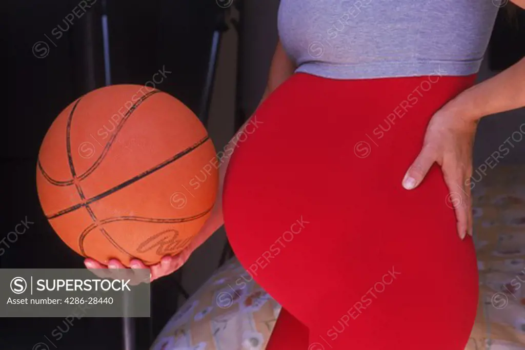 Pregnant lady during 8th month holding basketball next to equally round abdomen in bedroom