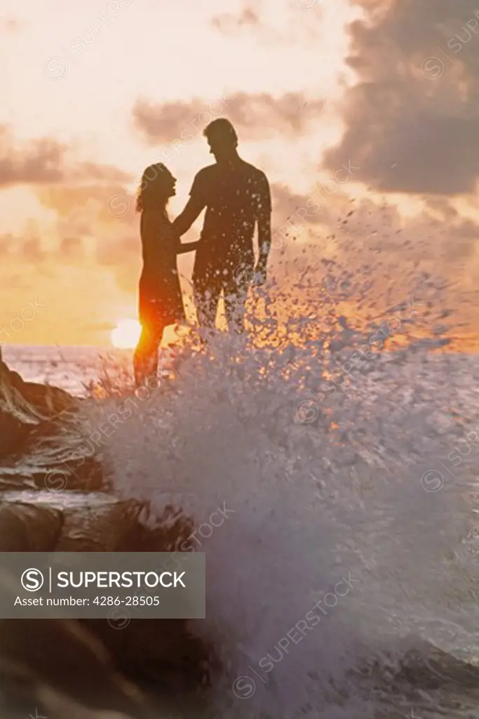 Couple standing on rocks at sunset with wave hitting shore in Seychelle Islands
