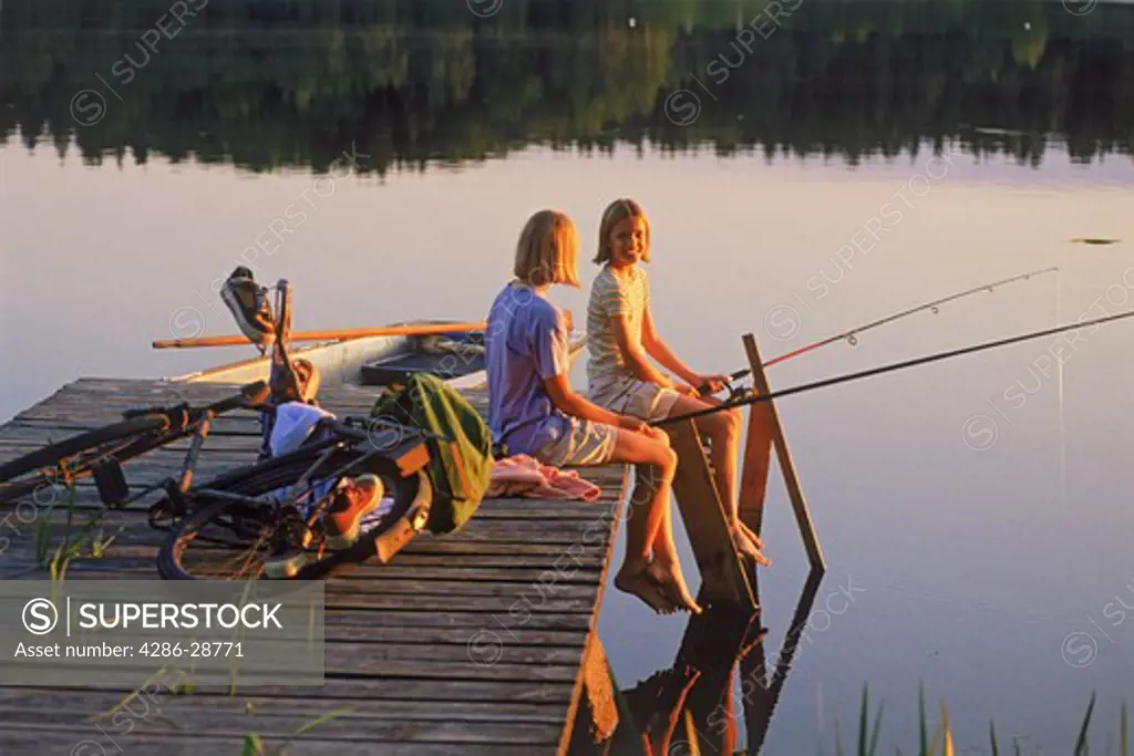Two girls 11 to 13 enjoying fishing picnic on lakeside pier in Sweden at sunset