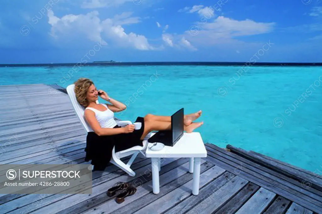 Woman with cellphone and laptop on beach chair and wooden dock in Maldives on working holiday