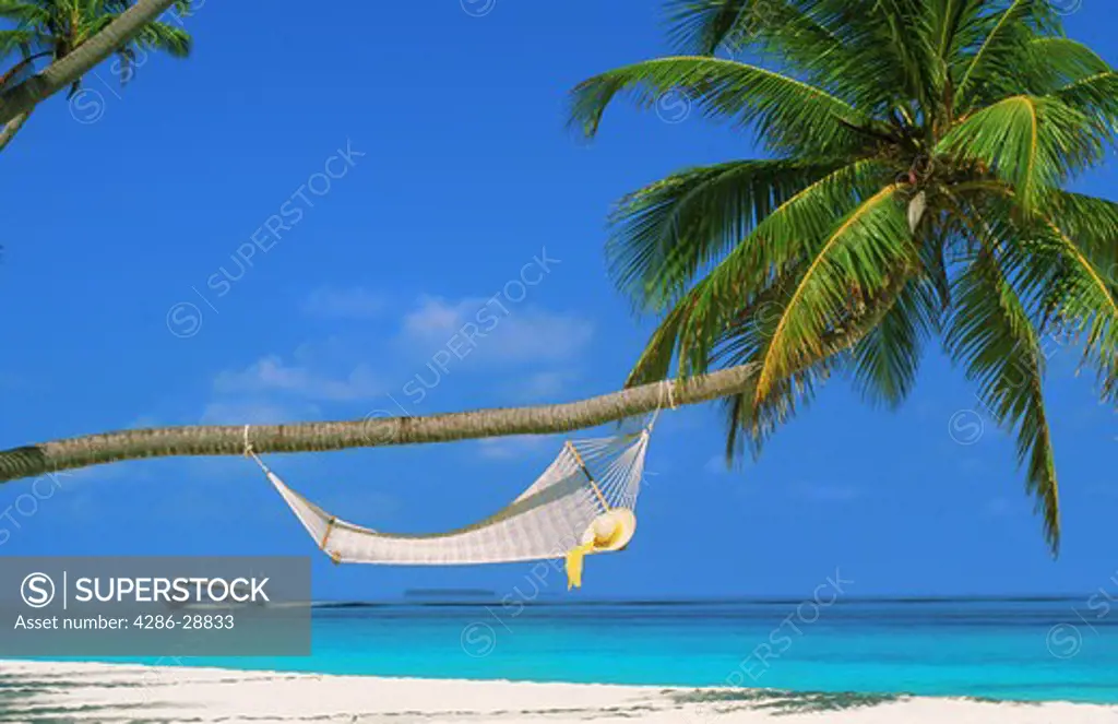 Hat resting on hammock over sandy shores in Maldive Islands with passing tourboat