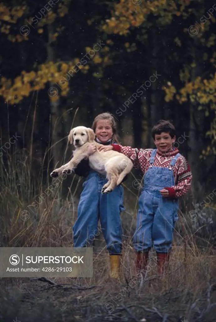 Happy, smiling boy and girl standing outside on a flurrying autumn day. The girl holds a golden retriever puppy in her arms.