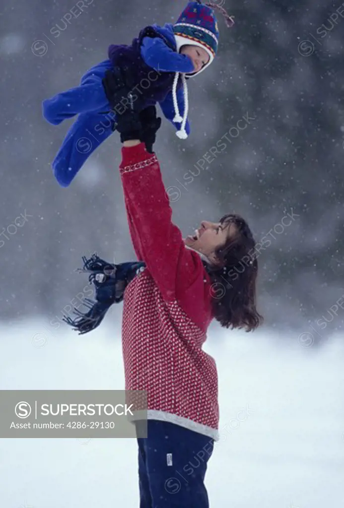 A mother lifts her baby high over her head while standing outdoors on a lightly snowing day.