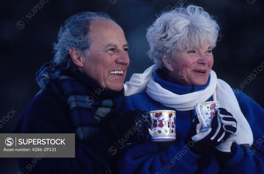 Portrait of a happy senior couple standing together outdoors on a chilly day and holding colorful mugs of a warm beverage.