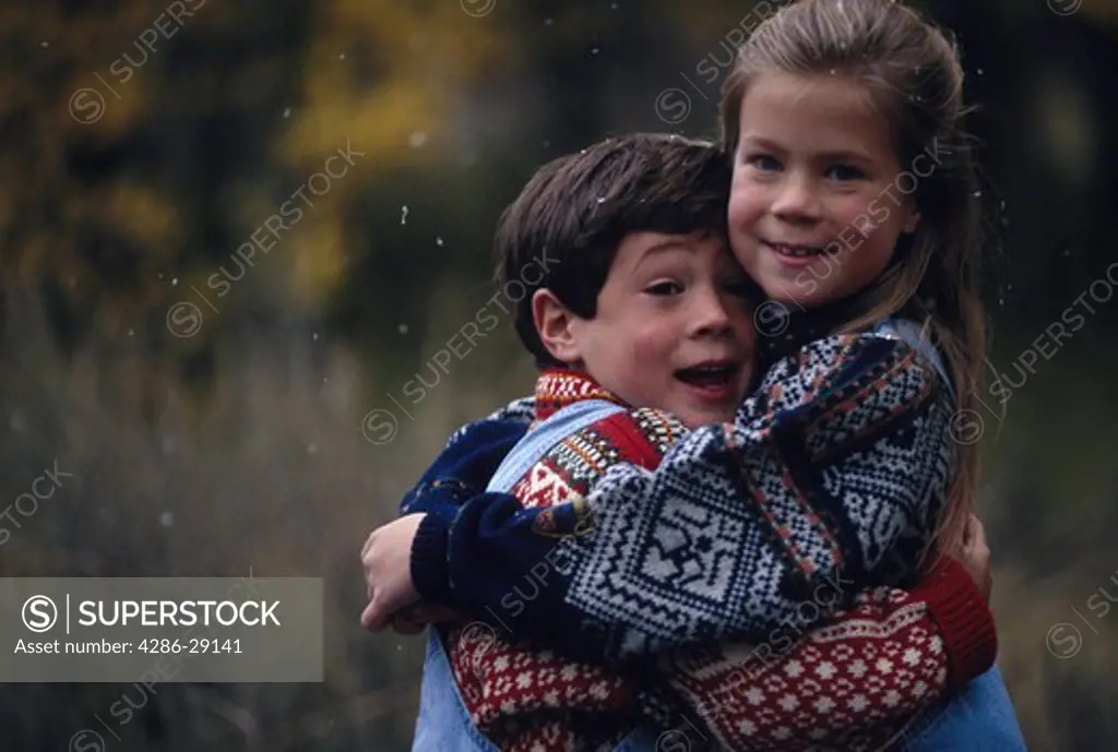 Portrait of a brother and sister hugging each other outdoors during a gentle snowfall.