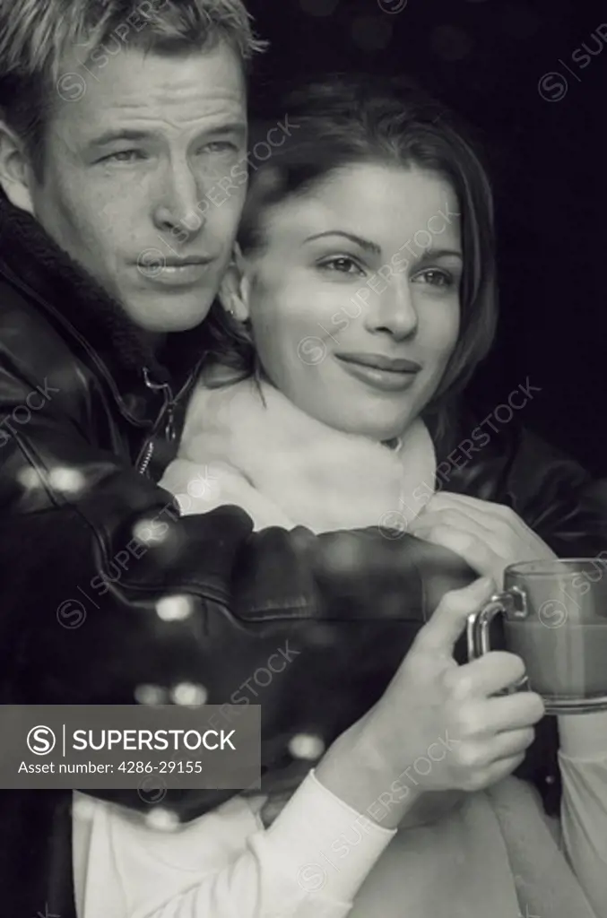 A man embraces a smiling woman as she holds a glass cup of coffee and looks off in the distance.