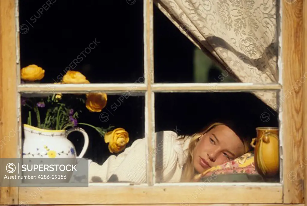 A young woman looks serenely out a rustic window as she rests on a pillow. 