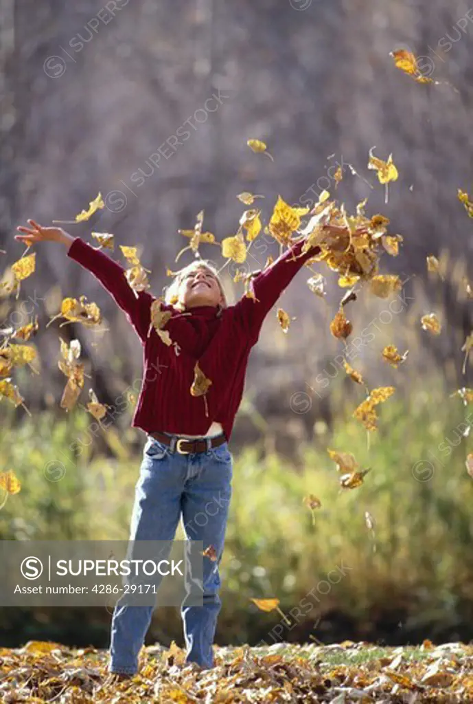 A young person happily tosses an armful of yellow leaves overhead on a sunny autumn day.