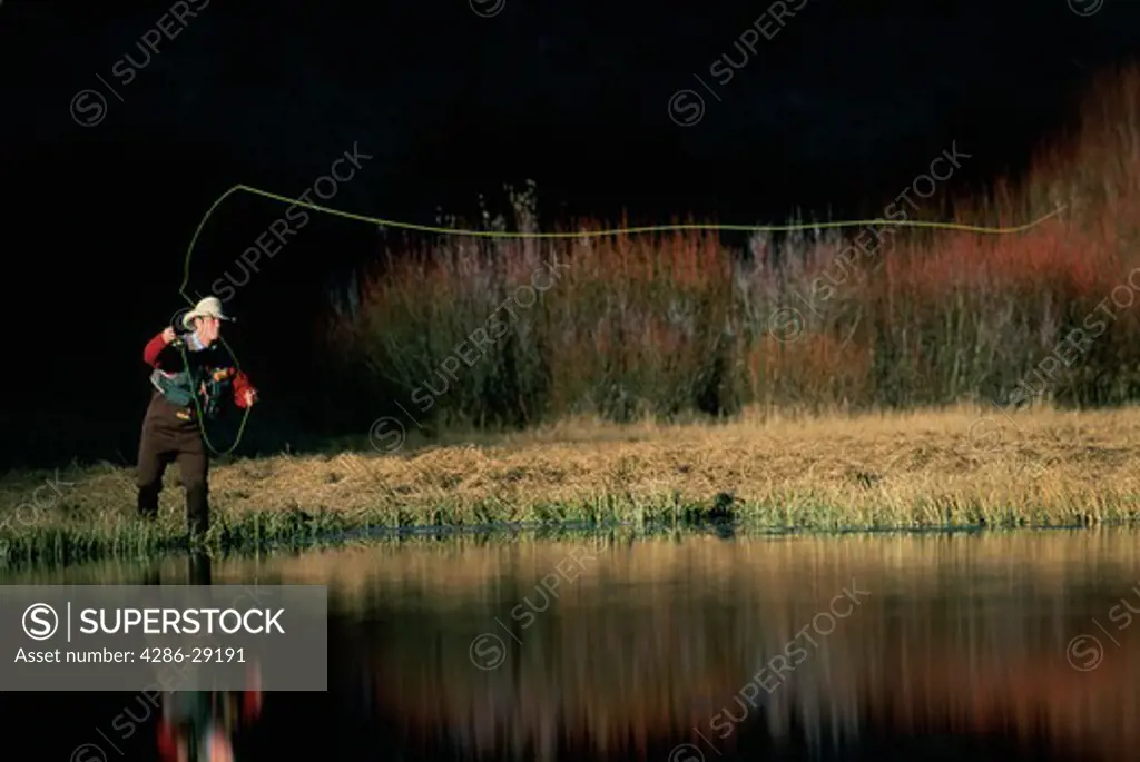 A man stands on shore and casts his fly-fishing line far across the still waters of a western lake.