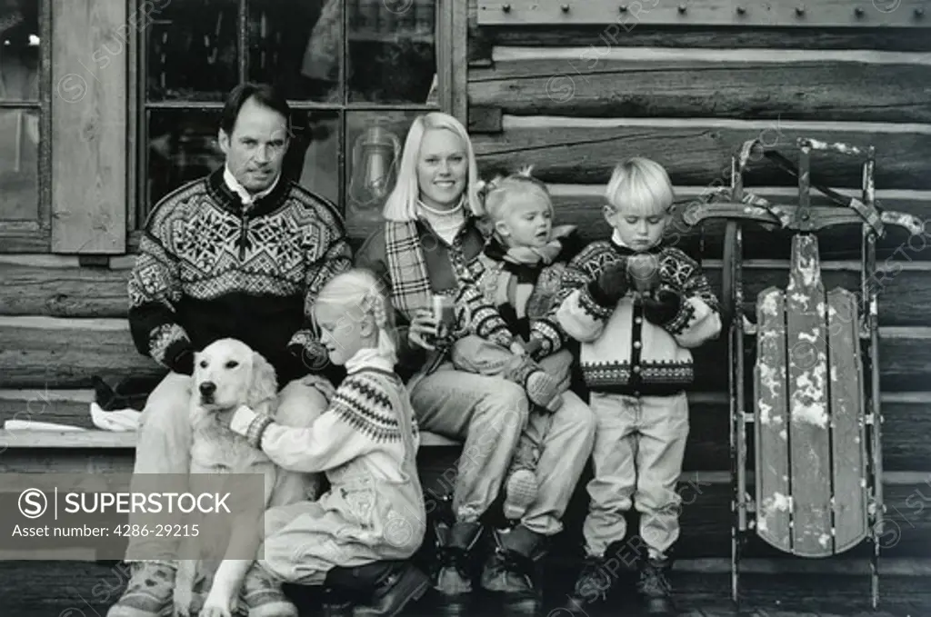 A couple and their three children together outside a log cabin on a chilly day drinking warm beverages and playing with their dog.