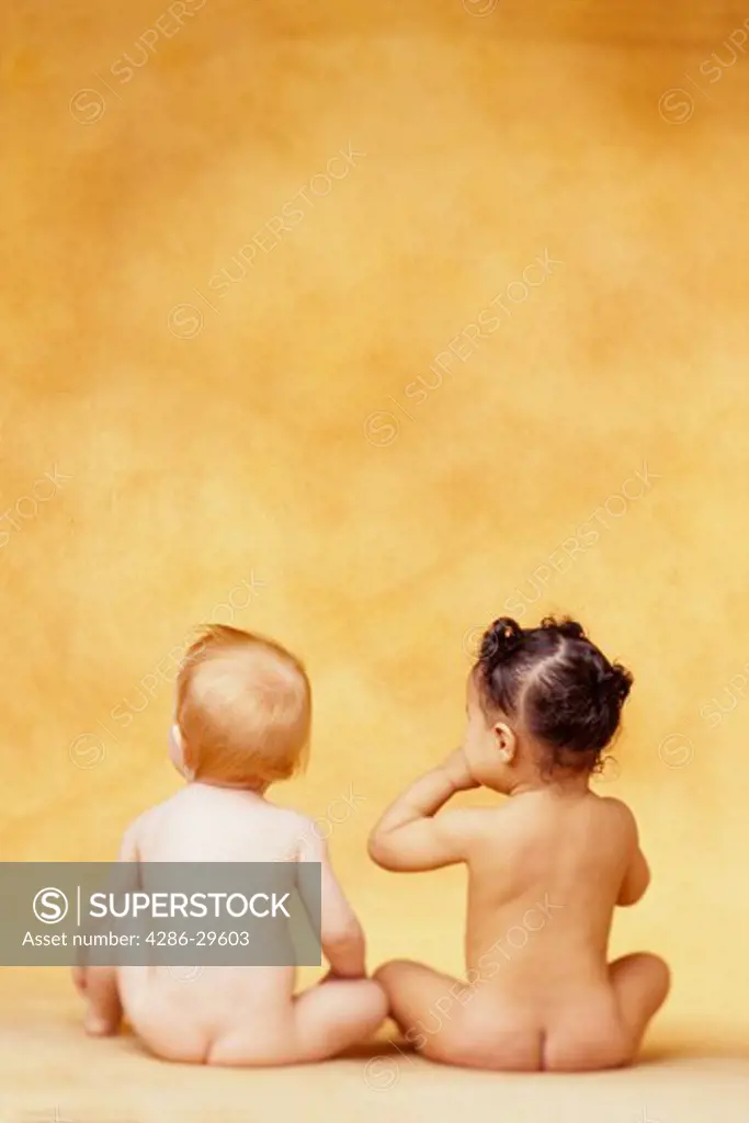 Two naked babies, one white and the other black, sit together in a studio with their backs to the camera.