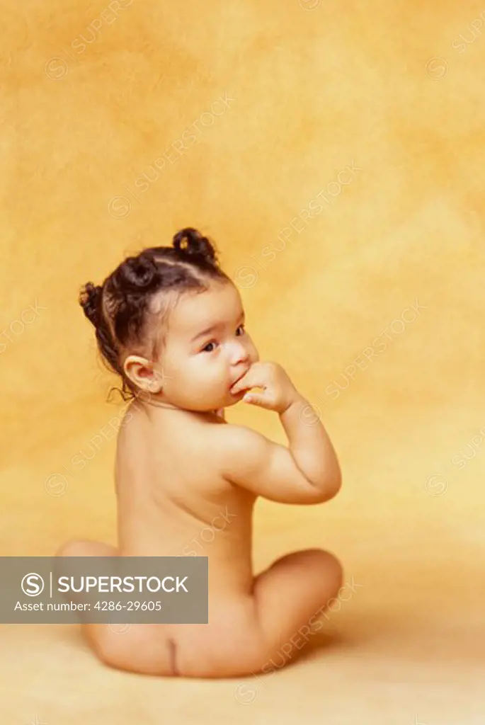 Naked African-American baby sits in a studio with her back to the camera and looks over her shoulder.