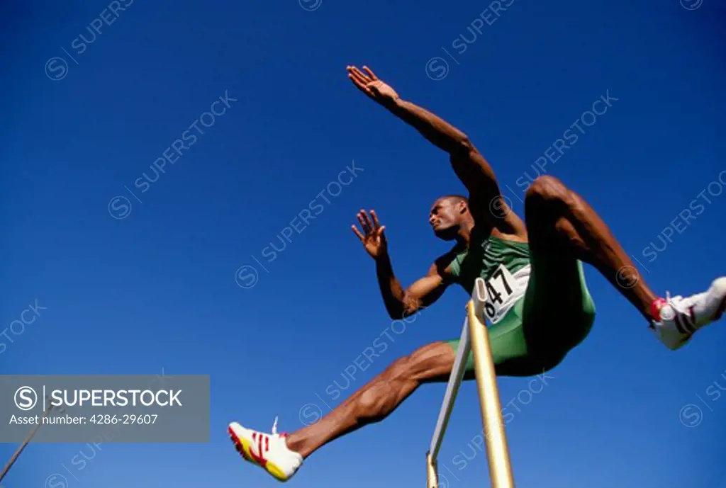 Looking upwards at a track runner as he leaps over a hurdle during a race.