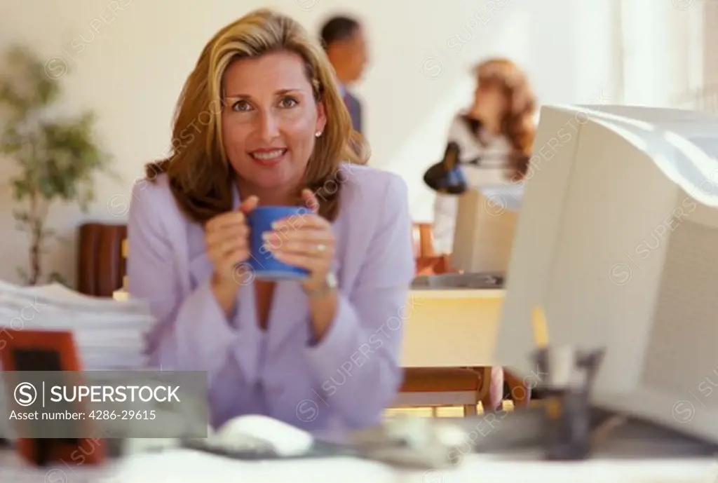 Female executive sits at her desk by her computer and holds a blue mug with both hands.